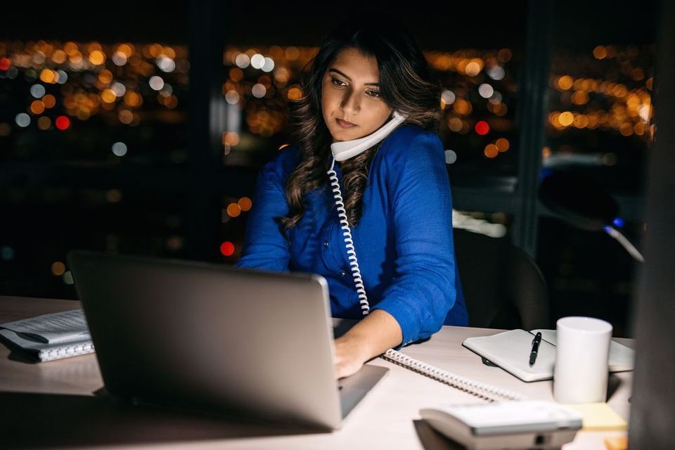 A woman sits in front of a laptop, holding a phone up to her ear with a shoulder.